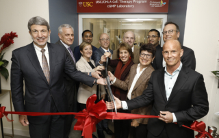 Eleven high-ranking professionals happily cut a red ribbon in front of a doorway with a sign reading, USC-CHLA Cell Therapy Program, CGMP Laboratory