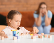 In a living room, a little girl sorts colored blocks as her mother looks on worriedly.