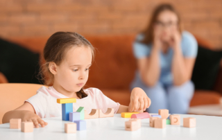 In a living room, a little girl sorts colored blocks as her mother looks on worriedly.