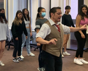 A trim, middle-aged man demonstrates a dance move for a small, attentive group of young people