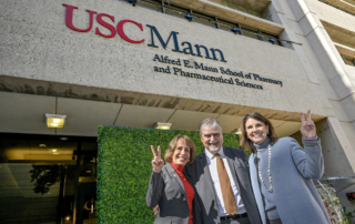 A man in a suit stands between two professionally dressed women in front of a facade reading USC Mann, Alfred E. Mann School of Pharmacy and Pharmaceutical Sciences