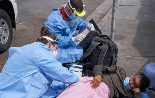 A woman and a man in full PPE give medical treatment to a man lying under a blanket on a filthy sidewalk