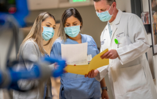 Three masked medical providers study a sheet of paper in a medical hallway