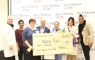 In front of a backdrop reading Keck Medicine of USC, a woman in blue scrubs stands between professionally dressed people holding a giant check