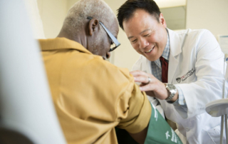 A smiling man in a white coat fits a blood pressure cuff around an older man's arm.