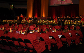 Graduates in cardinal commencement robes and mortarboards fill an auditorium, listening to a speaker onstage.
