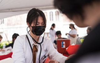 In a festival tent, a young woman in a white coat and medical mask tends to a member of the public.