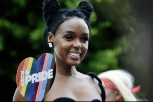 Cultural icon Janelle Monáe beams as she holds a paper fan that reads Keck Pride against a rainbow backdrop.