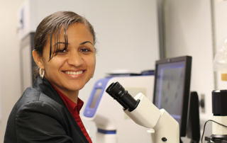 Senta Georgia, PhD, sits at a microscope and grins at the camera