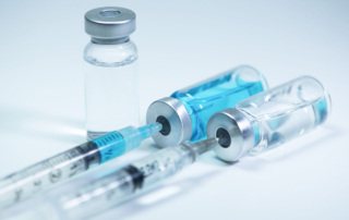 Two syringes with hypodermic needles each extract clear, blue fluid from a vial.