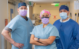 Three people in scrubs, surgical caps and masks stand proudly in a hospital hallway.