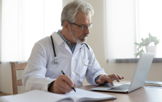 A bespectacled doctor with a gray beard studies at a laptop