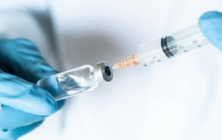 A closeup shows gloved hands using a syringe to extract clear fluid from a vial.