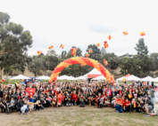 A large group of people in matching cardinal T-shirts pose for a photo in front of a ballon arch in cardinal and gold.
