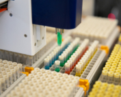 In a lab, a machine with a small, green attachment hovers over a rack of multicolored test tubes