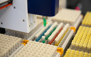 In a lab, a machine with a small, green attachment hovers over a rack of multicolored test tubes