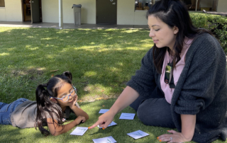 On a grassy lawn, a young woman teaches letters to a little girl.