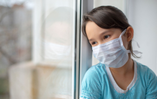 A little girl in a face mask gazes out a window.