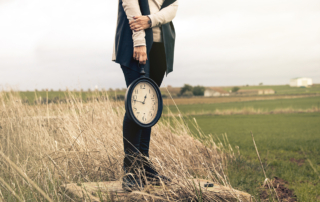 Beneath an overcast sky, a woman holds a wall clock as she stands in a field of patchy grass.