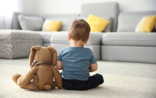 A little boy sits alone on the floor with a plush toy, his back to the camera.