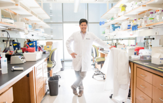 A trim man in a white coat smiles brightly in an extensive research lab