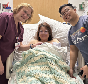 A woman in a hospital bed cradles a newborn beneath a hand-knitted baby blanket